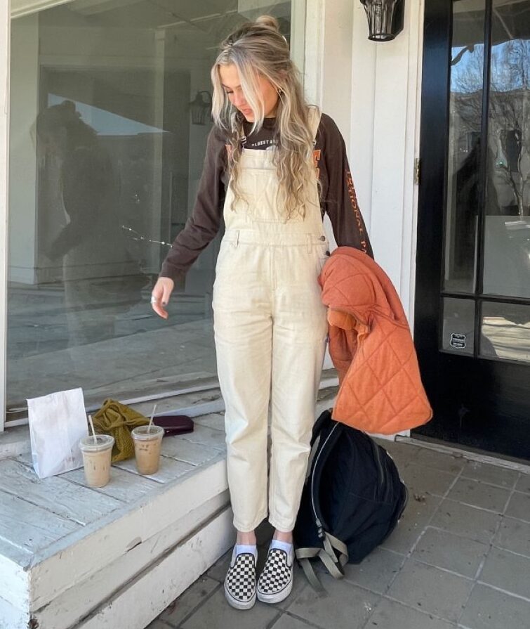 blonde woman wearing cream overalls with a long sleeve shirt underneath and checkered vans, drinking iced coffee.