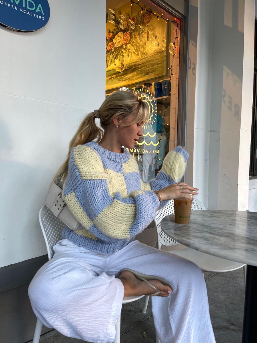 blonde woman sitting down, drinking an iced coffee, wearing a checkered blue and cream sweater and flip flops