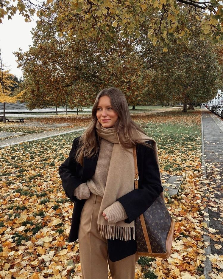 woman in a pile of leaves, wearing a louis vuitton bag, brown pleated pants, brown sweater, brown cashmere scarf, and black blazer