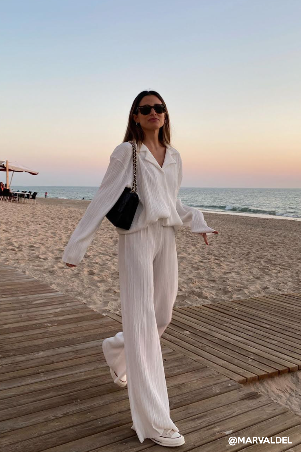 brunette woman walking on the beach, wearing an all cream long sleeve pant set loungewear and black chanel bag.