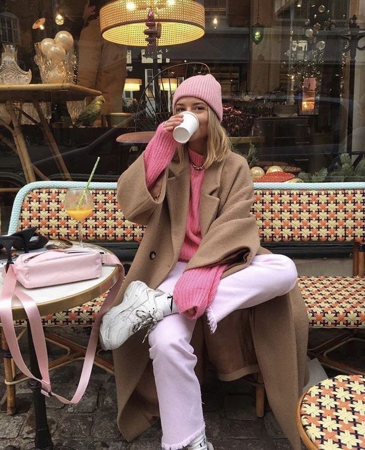 woman sitting outside on a patterned bench drinking a coffee, wearing an all pink outfit and brown long coat.