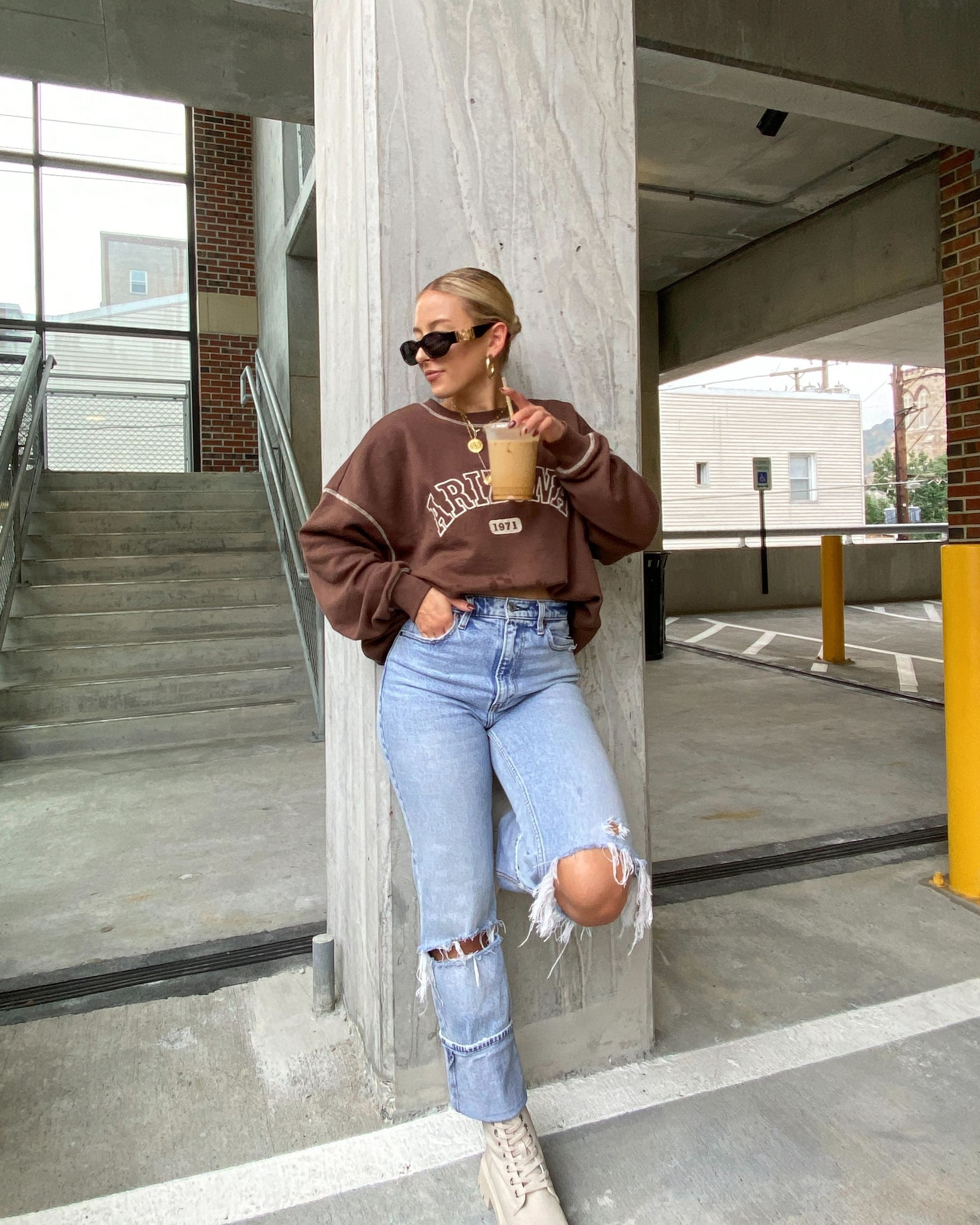 blonde woman wearing ripped jeans and a brown Arizona sweatshirt drinking an iced coffee.