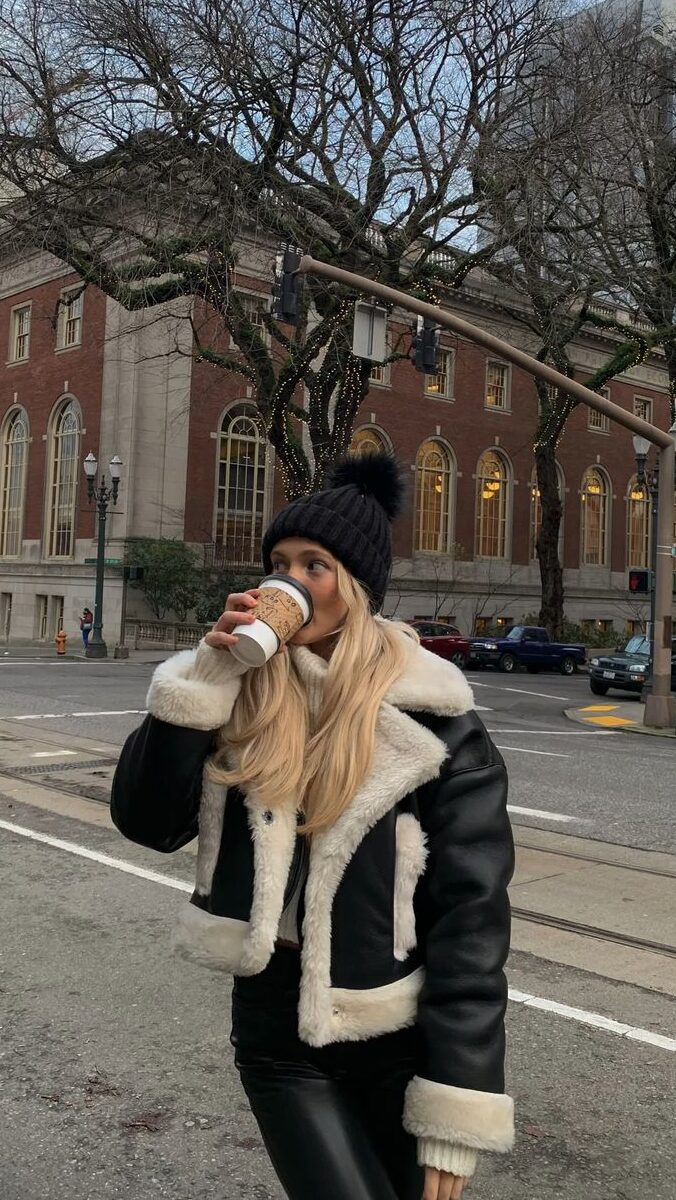blonde woman in big city wearing a black leather jacket with fur on the inside, drinking a cup of coffee and wearing a black beanie.