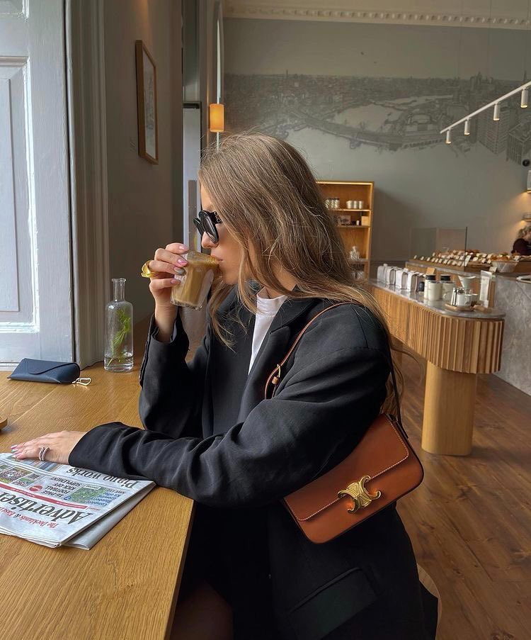woman in coffee shop reading a newspaper and drinking a coffee, wearing a black blazer and brown bag